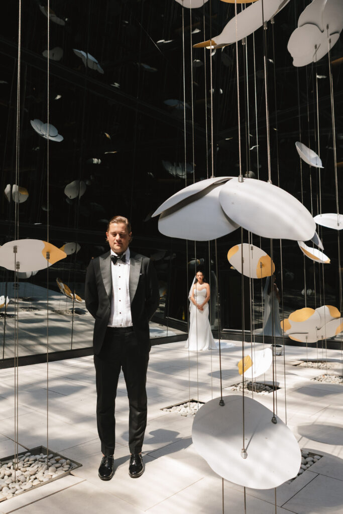 Modern wedding portrait of groom in black tuxedo with bride in white gown framed by hanging sculptural installation at Four Seasons Montreal