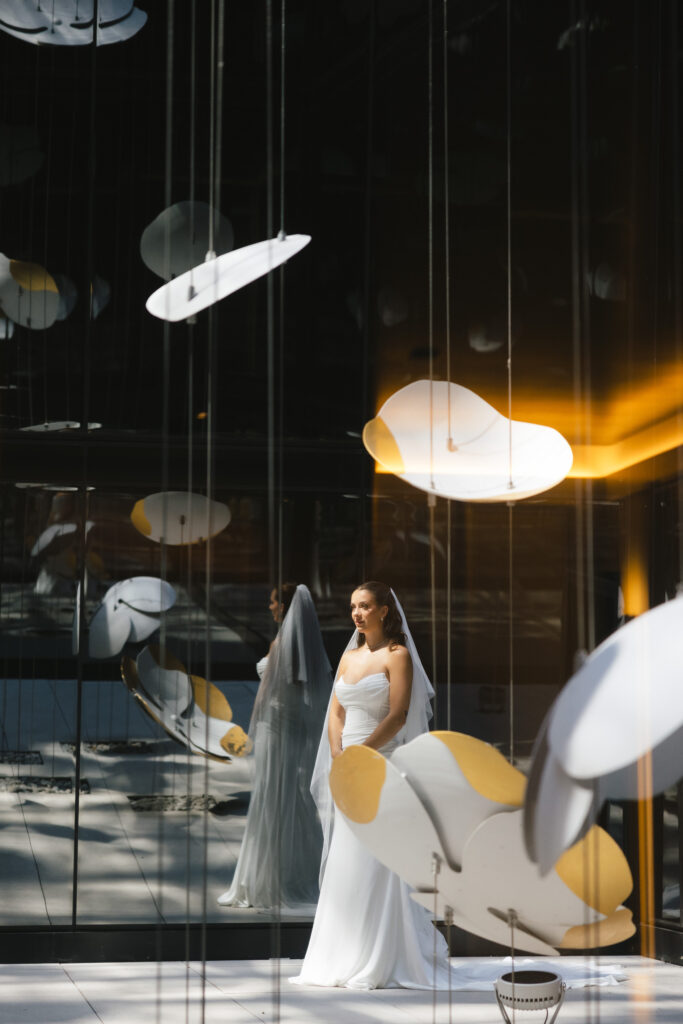 Bride of Samuel Montembeault in strapless wedding gown and veil standing beneath modern hanging art installation at contemporary wedding venue of Four Seasons Hotel