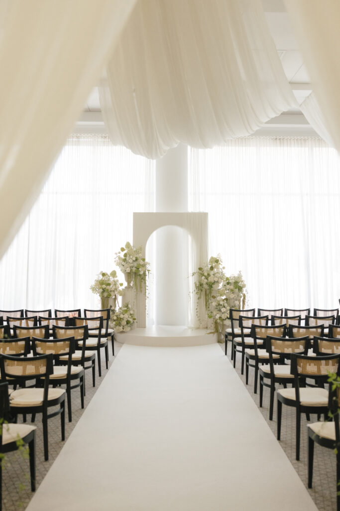 Elegant indoor wedding ceremony setup with white aisle runner, floral arch altar, and soft draped fabric in a light-filled venue