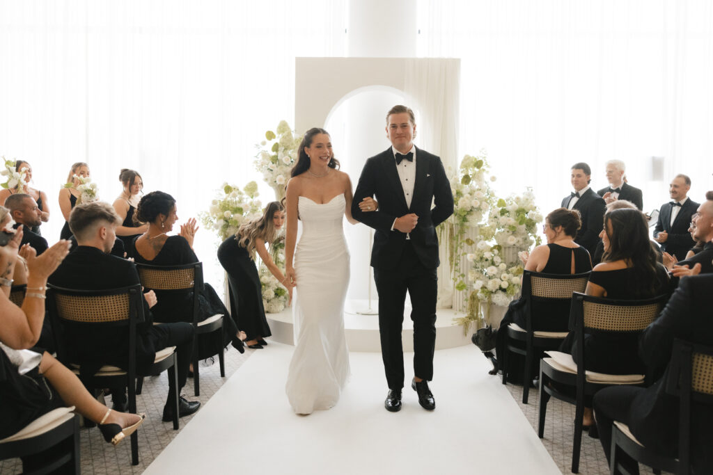 Newlyweds walking down the aisle after their indoor wedding ceremony in Four Seasons Montreal, surrounded by guests and white floral arrangements from Atelier Carmel