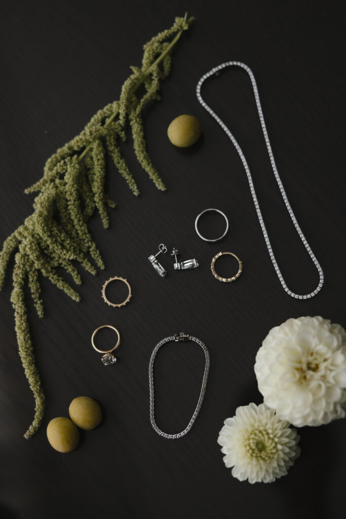 Flat lay of wedding jewelry including gold wedding bands, diamond engagement ring, earrings, and necklace styled with white florals and greenery on a dark background