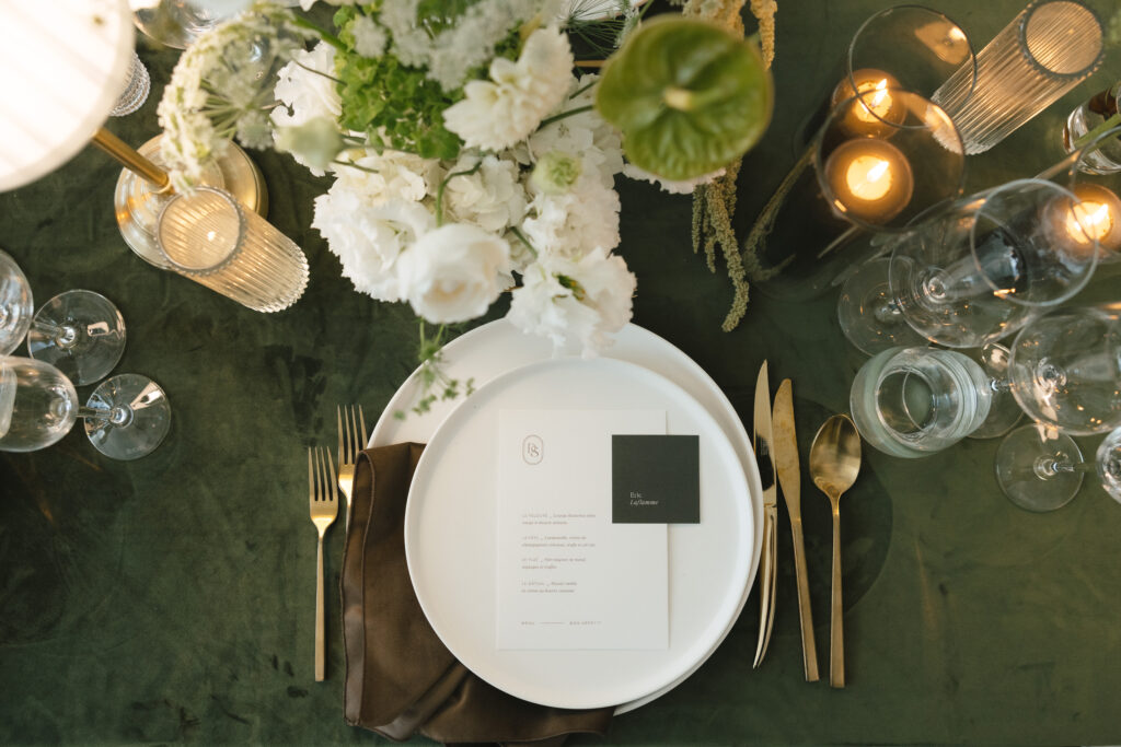 Elegant wedding table setting with white place card, layered dinnerware, gold flatware, glassware, candles, and floral centerpiece styled on a green tablecloth styled by La Vie Est Une Fête