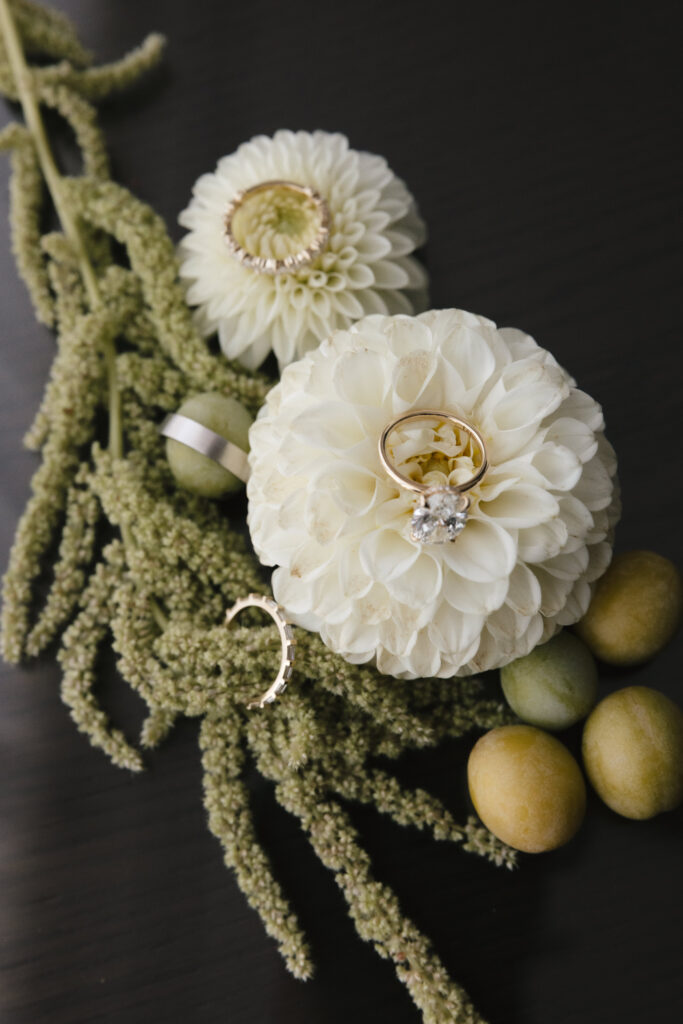 Wedding detail flat lay featuring white floral ring holders with gold wedding bands and diamond engagement ring on dark surface.