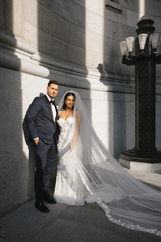 Bride and groom standing together in the soft sunlight, showcasing the bride's intricate wedding gown and flowing veil against the backdrop of elegant architecture