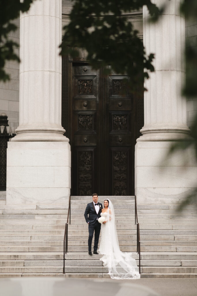 Bride and groom standing together on the grand steps of a historic building, showcasing their wedding attire and the architectural beauty of the Mary Queen of the World Cathedral