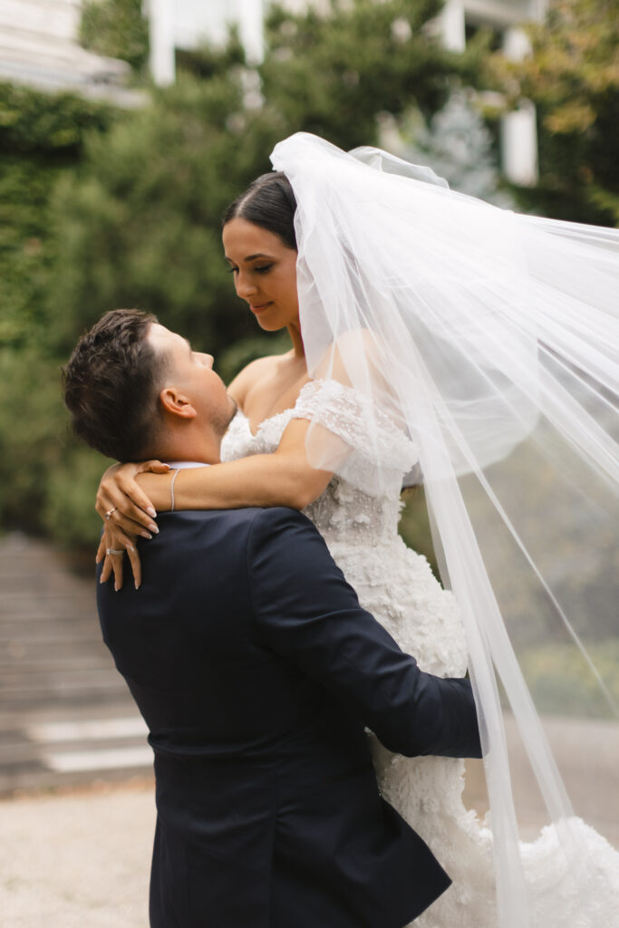 Bride and groom sharing an intimate moment, with the groom lifting the bride, showcasing her stunning wedding gown and flowing veil in a romantic outdoor setting