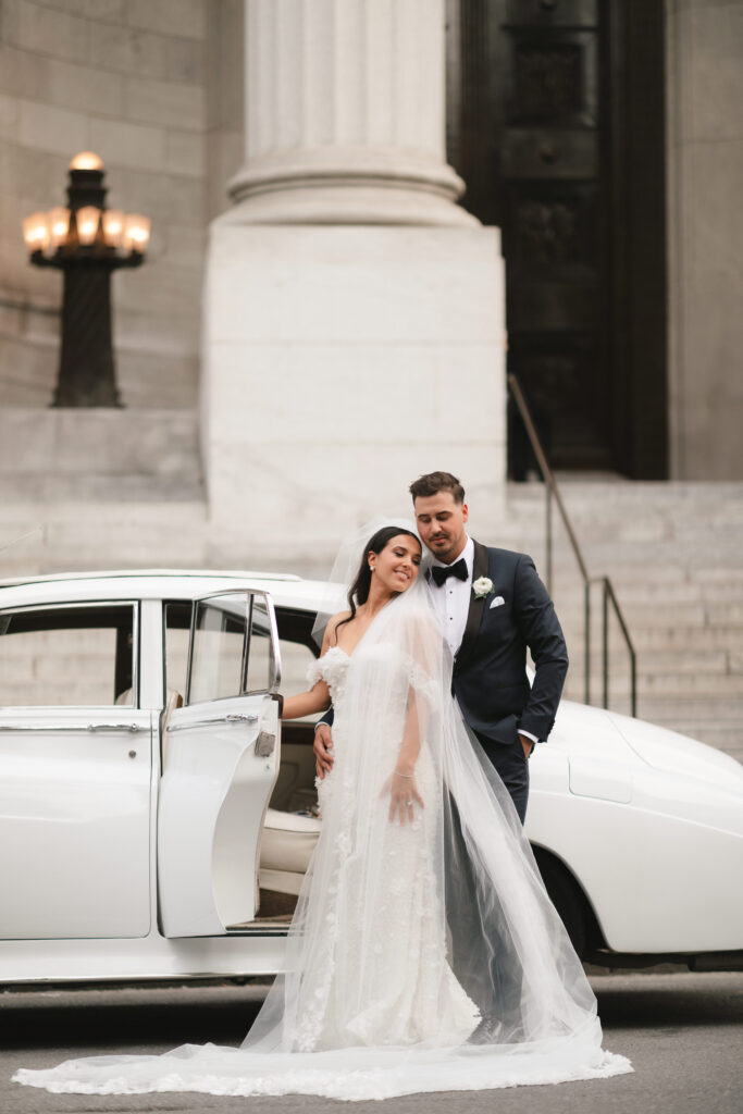Bride and groom standing in front of a classic white wedding car, with the bride in a stunning gown and long veil, capturing a moment of elegance at the historic Maison Principale