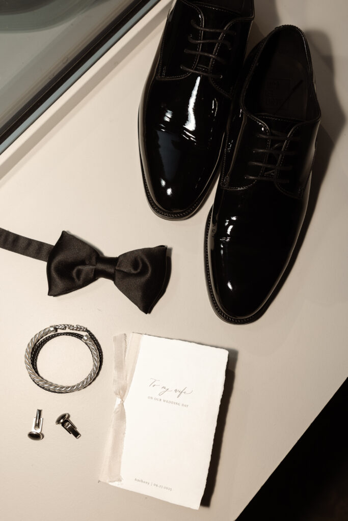 Groom’s wedding attire details including black polished shoes, bow tie, bracelet, cufflinks, and wedding invitation
